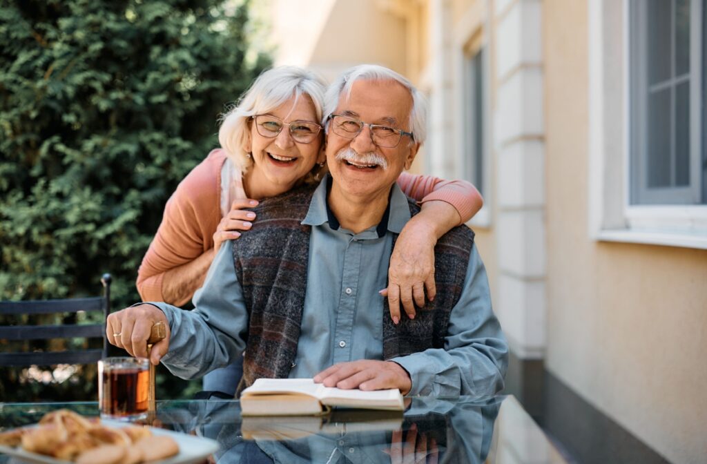 An older married couple smiling outdoors at the camera in a beautiful common space of a senior living community.